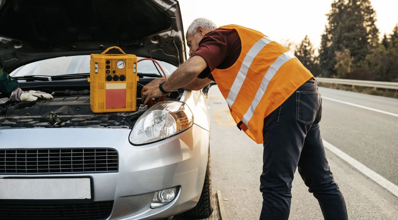 Roadside assistance technician in orange safety vest jump-starting a car battery on a highway