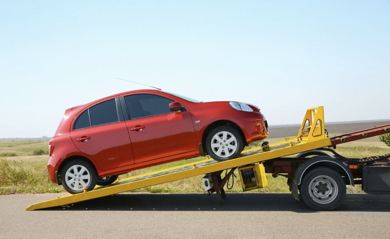 Red compact car being loaded onto a yellow flatbed tow truck on a rural highway