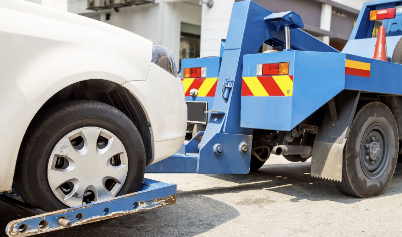 Blue wheel-lift tow truck assisting a stranded white car in need of battery service in Amityville, NY